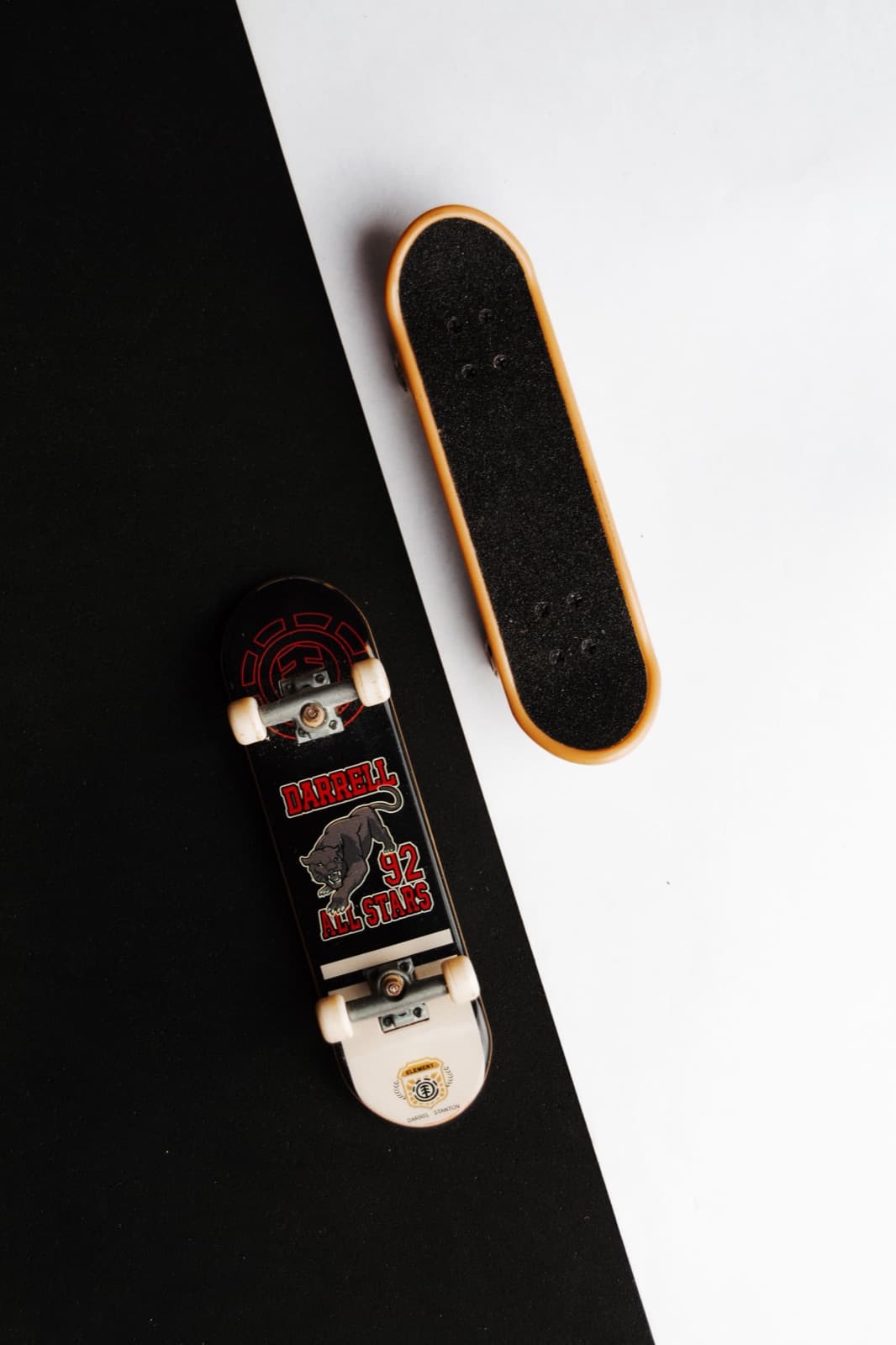Two miniature fingerboard skateboards seen from above on a black-and-white surface.